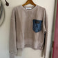 Brown sweatshirt with a blue pocket on a wooden hanger against a white wall.