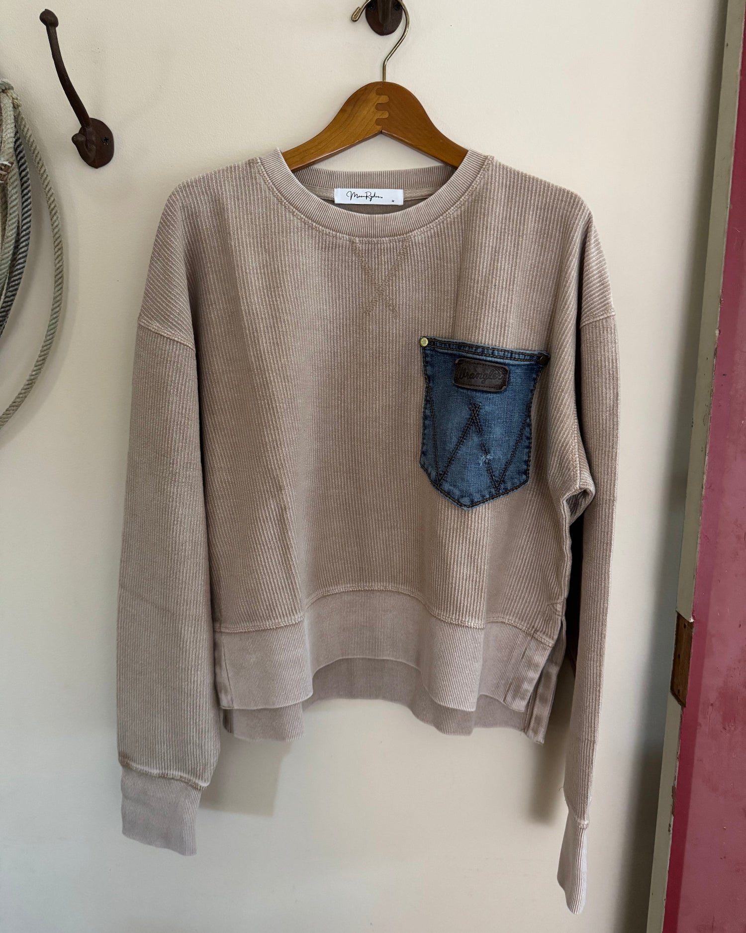 Brown sweatshirt with a blue pocket on a wooden hanger against a white wall.