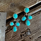 A pair of silver drop earrings with multiple turquoise-colored stones, displayed on a rustic wooden surface next to a metal ruler for scale.