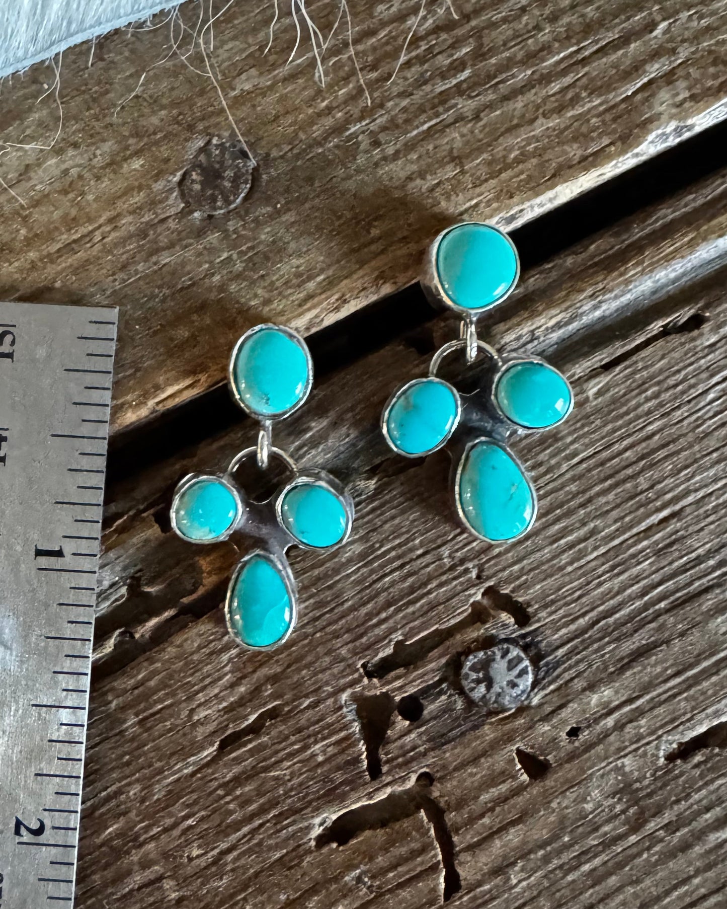 A pair of silver drop earrings with multiple turquoise-colored stones, displayed on a rustic wooden surface next to a metal ruler for scale.