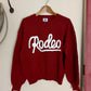 Red sweater with 'Rodeo' text on a hanger against a white wall.