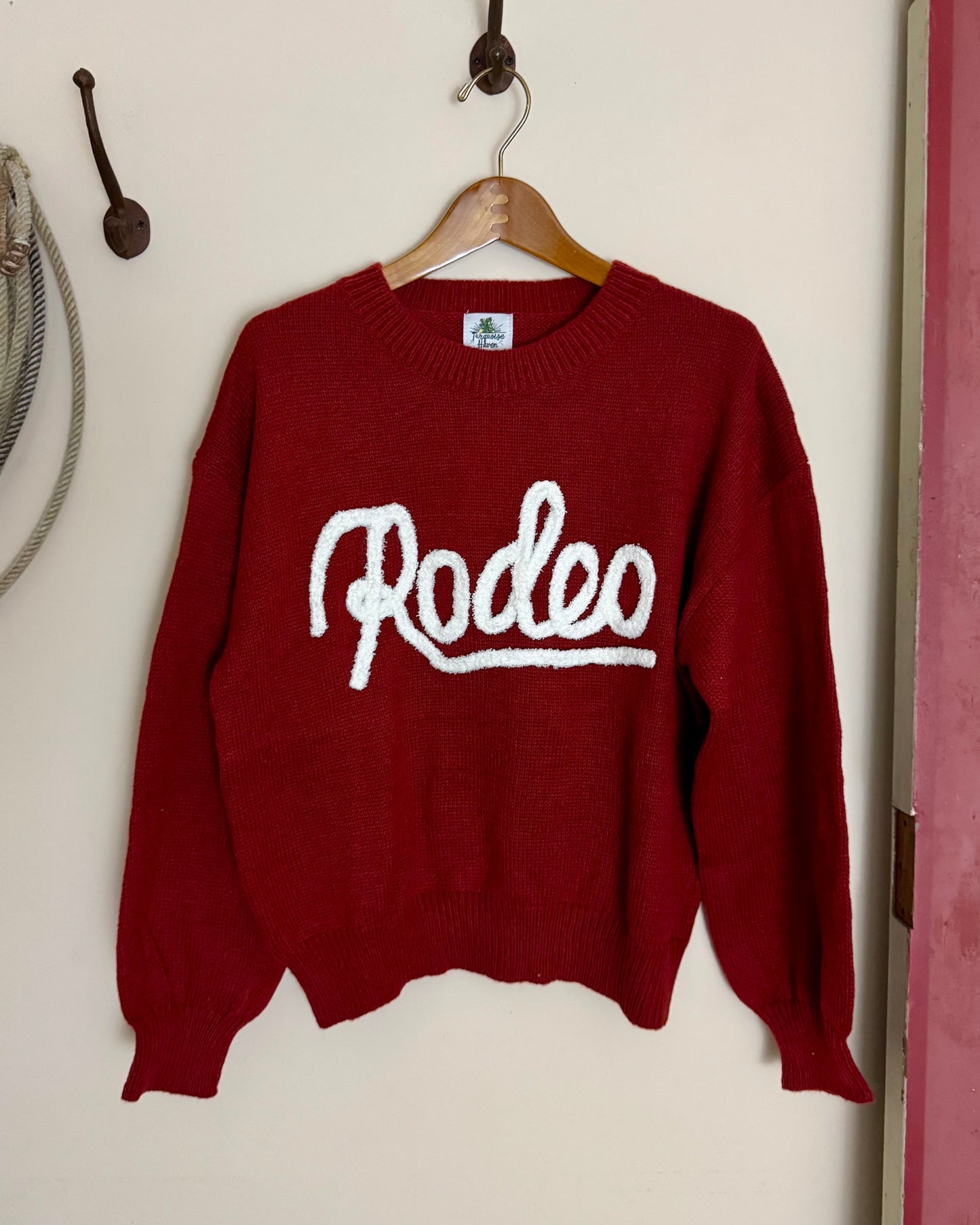 Red sweater with 'Rodeo' text on a hanger against a white wall.