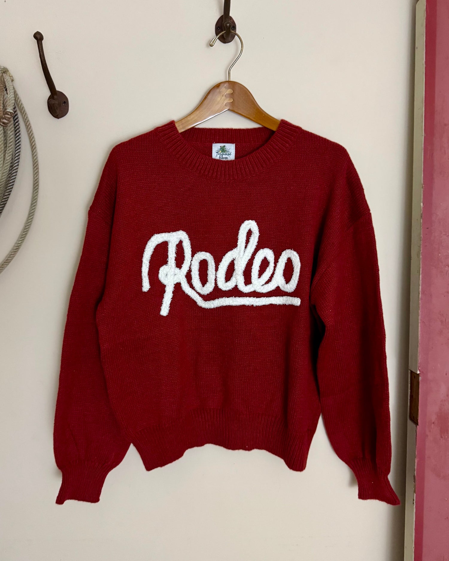 Red sweater with 'Rodeo' text on a hanger against a white wall.