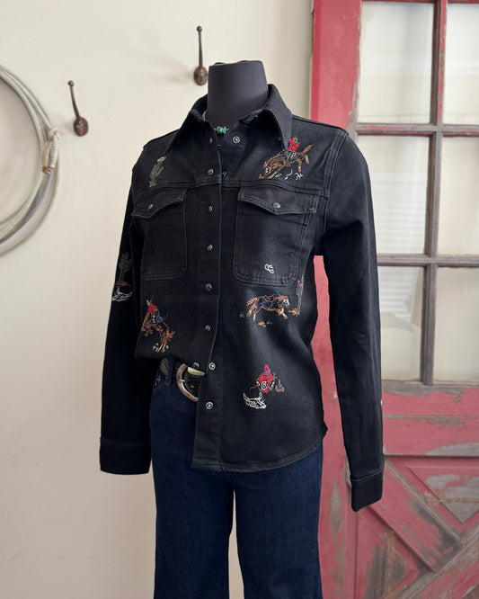 Black denim jacket with cowboy embroidery on a mannequin against a rustic background.