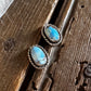 A pair of silver-toned stud earrings with oval turquoise-blue stones, displayed on a rustic dark wood surface next to white fur.