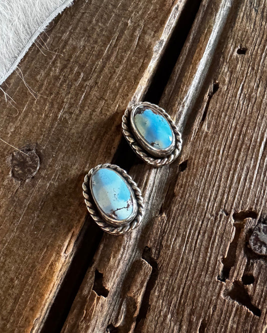 A pair of silver-toned stud earrings with oval turquoise-blue stones, displayed on a rustic dark wood surface next to white fur.