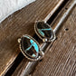 A pair of oval turquoise and black stone earrings in a twisted silver-tone setting, resting on a rustic wooden surface with a white faux-fur textile in the background.