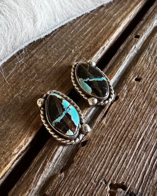 A pair of oval turquoise and black stone earrings in a twisted silver-tone setting, resting on a rustic wooden surface with a white faux-fur textile in the background.