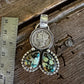 A sterling silver pendant featuring a coin-set oval and three marquise-shaped green iridescent stones, displayed on a rustic wooden surface next to a metal ruler for scale.