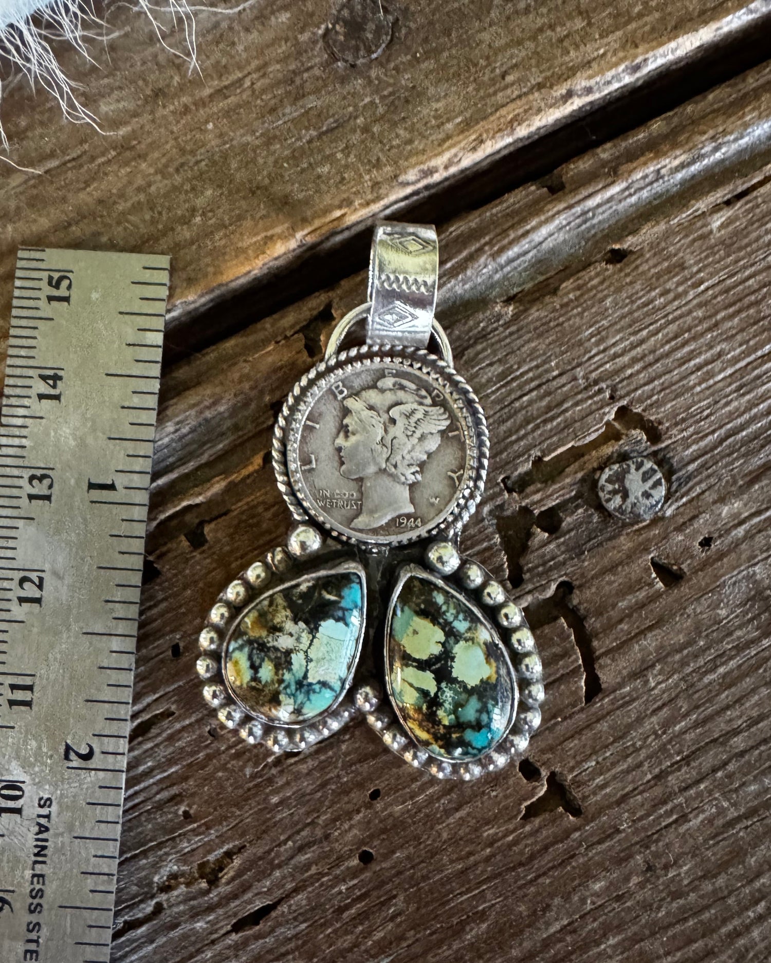 A sterling silver pendant featuring a coin-set oval and three marquise-shaped green iridescent stones, displayed on a rustic wooden surface next to a metal ruler for scale.