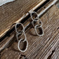 A pair of silver-toned dangle earrings with three interlocking textured oval links, displayed on a rustic dark wood surface with a white faux-fur fabric at the top.