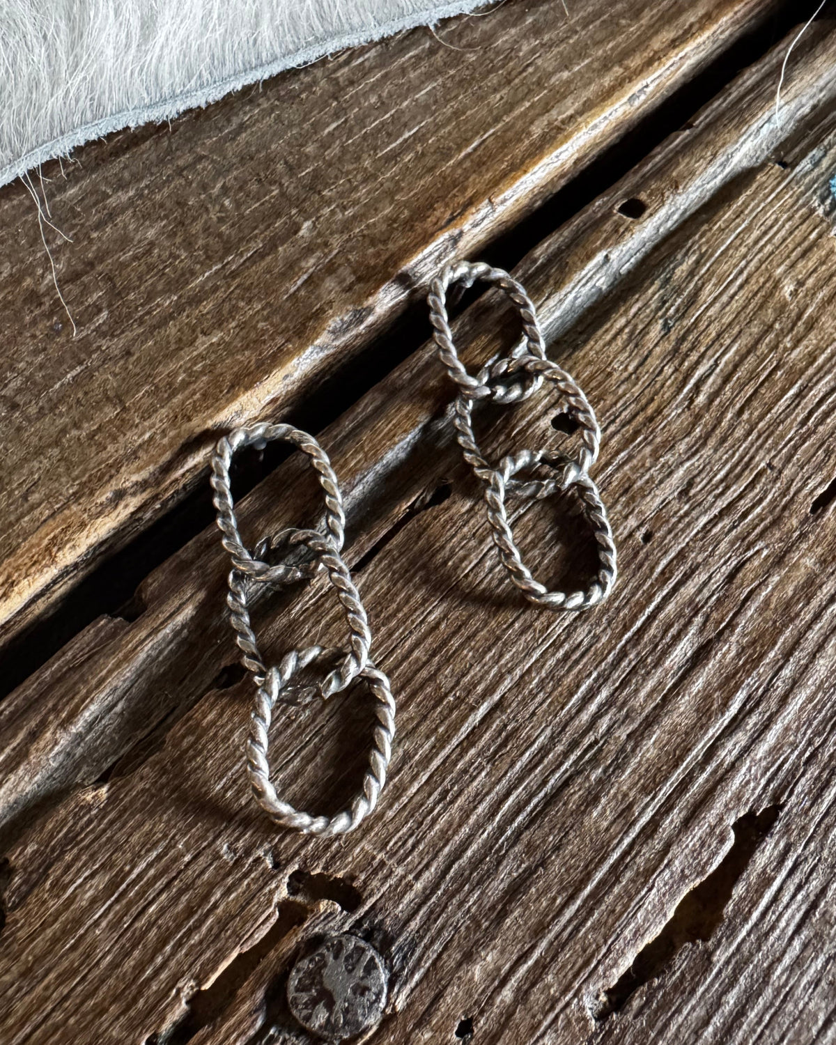 A pair of silver-toned dangle earrings with three interlocking textured oval links, displayed on a rustic dark wood surface with a white faux-fur fabric at the top.
