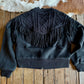 Black sweater with fringe details on a wooden surface with a cowhide rug in the background