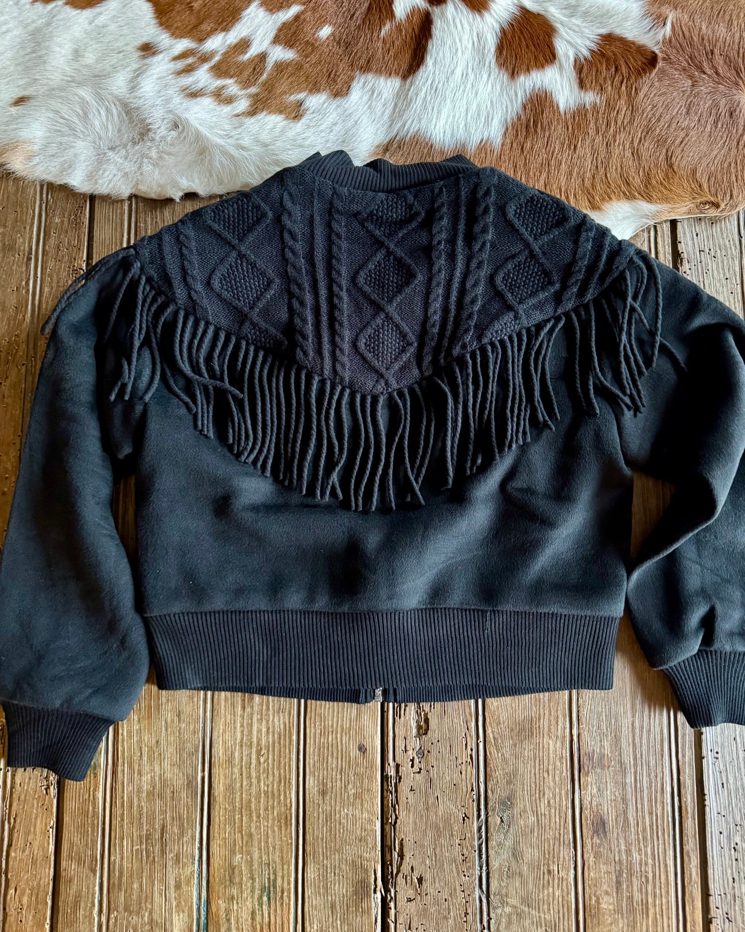 Black sweater with fringe details on a wooden surface with a cowhide rug in the background