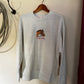 A light gray heather crewneck sweatshirt hanging on a wooden hanger from a wall hook. The sweatshirt features a small graphic on the left chest of a brown horse with red text underneath that reads 'and by Goddy'.
