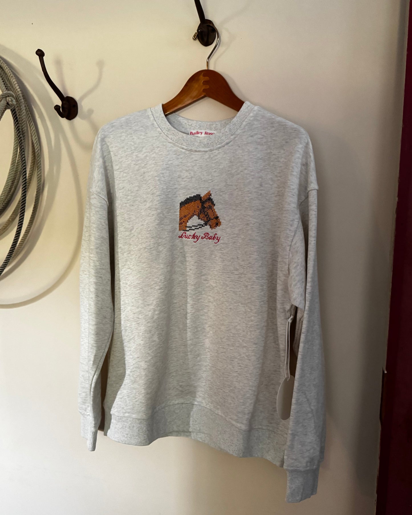 A light gray heather crewneck sweatshirt hanging on a wooden hanger from a wall hook. The sweatshirt features a small graphic on the left chest of a brown horse with red text underneath that reads 'and by Goddy'.