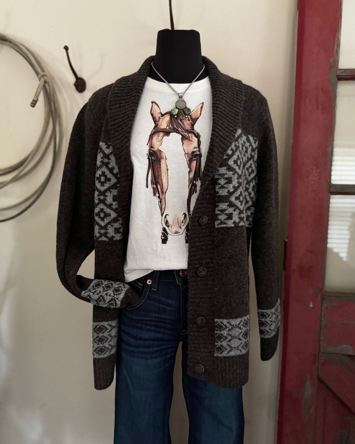 A dark brown knit cardigan with gray geometric patterns on the sleeves and hem, styled on a mannequin over a white graphic t-shirt and blue jeans.