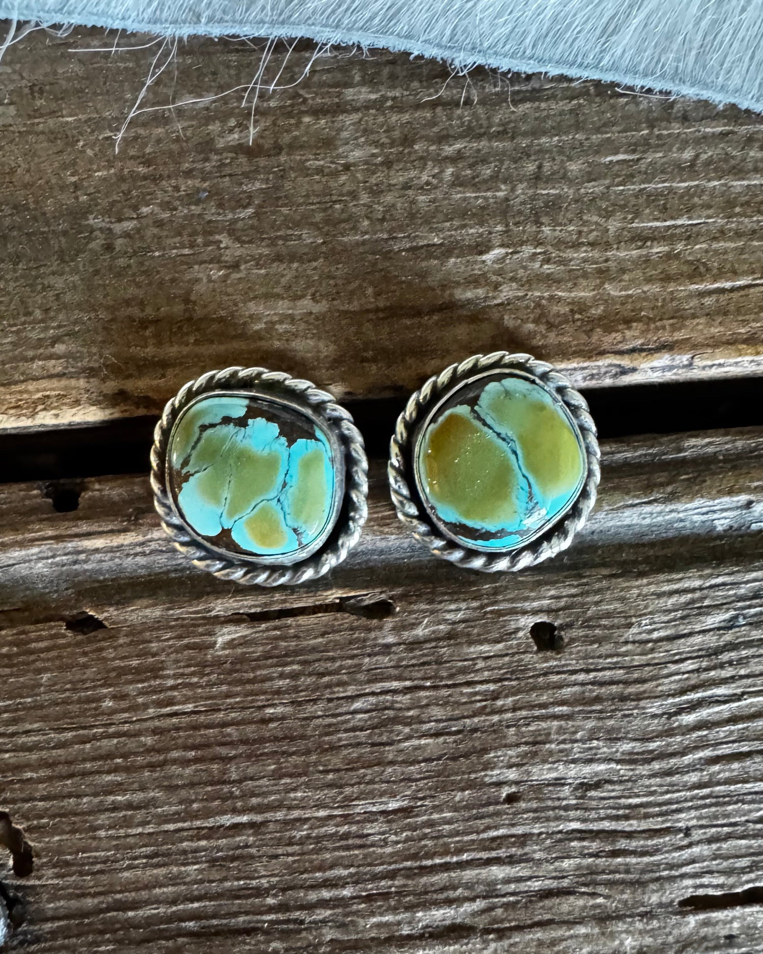 A pair of oval stud earrings with turquoise-blue and yellow marbled stones in a twisted silver-tone metal setting, displayed on a rustic wooden surface.