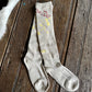 A pair of ribbed beige crew socks with yellow and red graphic details, laid flat on a dark wooden plank surface.