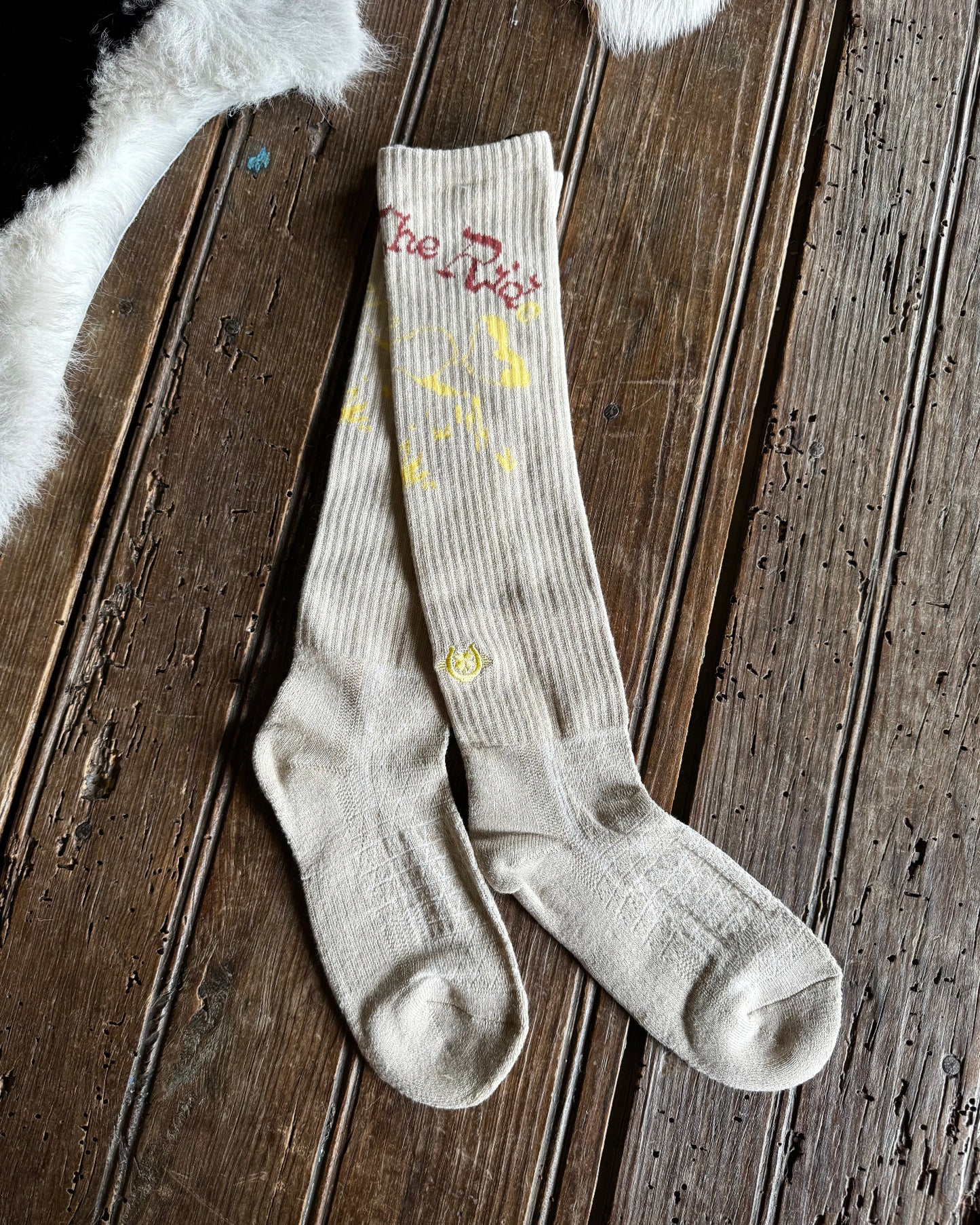 A pair of ribbed beige crew socks with yellow and red graphic details, laid flat on a dark wooden plank surface.