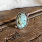 A sterling silver ring with a large oval turquoise stone, displayed on a rustic wooden surface with a white faux fur trim in the background.