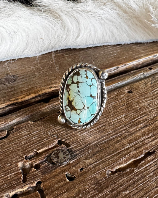 A sterling silver ring with a large oval turquoise stone, displayed on a rustic wooden surface with a white faux fur trim in the background.