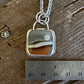 A silver necklace with a square pendant featuring a band of agate stone in earthy brown, beige, and gray tones, displayed on a rustic wooden surface next to a metal ruler for scale.