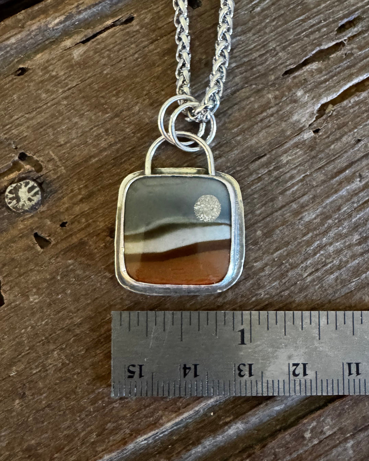 A silver necklace with a square pendant featuring a band of agate stone in earthy brown, beige, and gray tones, displayed on a rustic wooden surface next to a metal ruler for scale.
