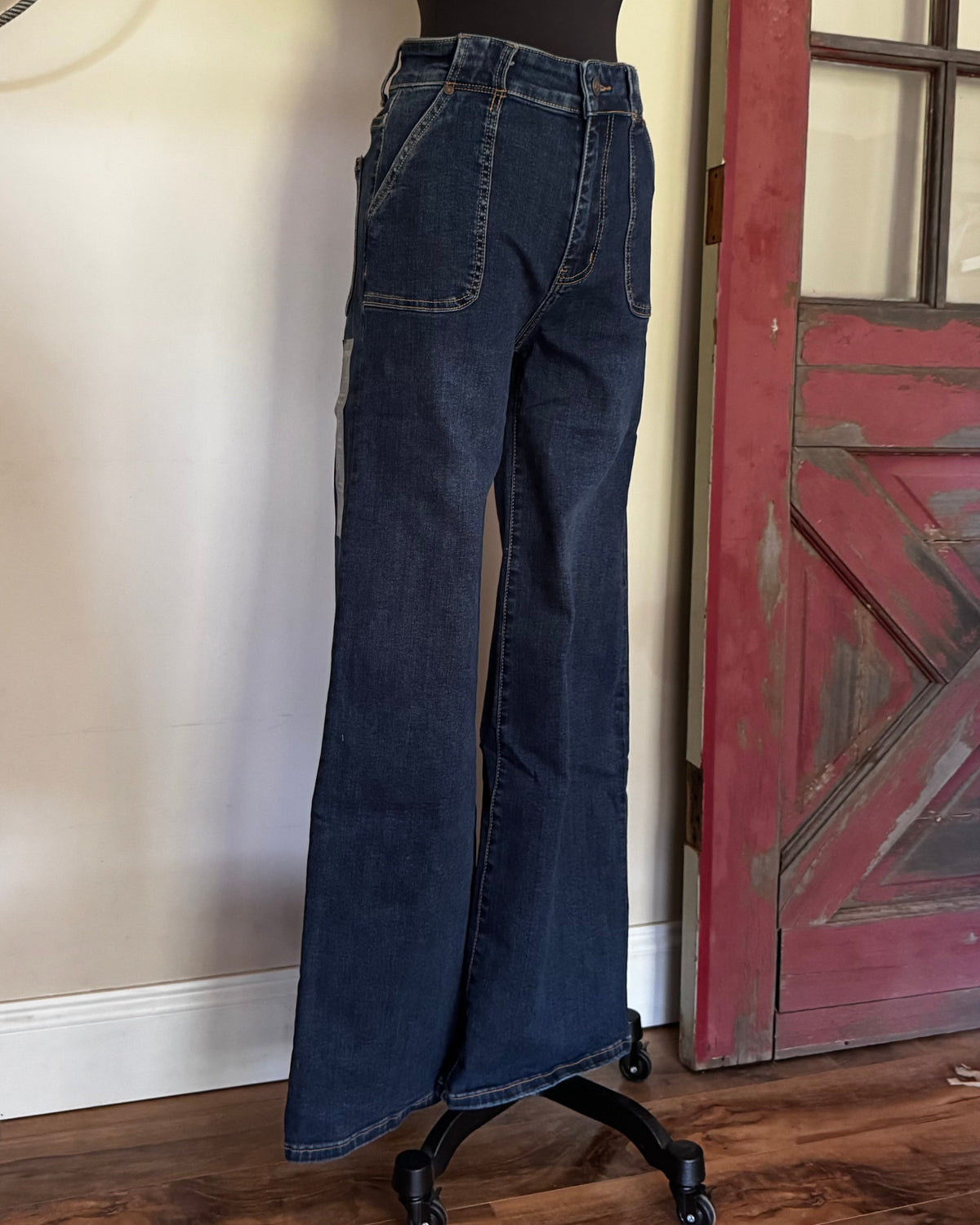 Dark blue wide-leg flare jeans displayed on a black dress form mannequin in an indoor setting with a wooden floor and a red distressed door.