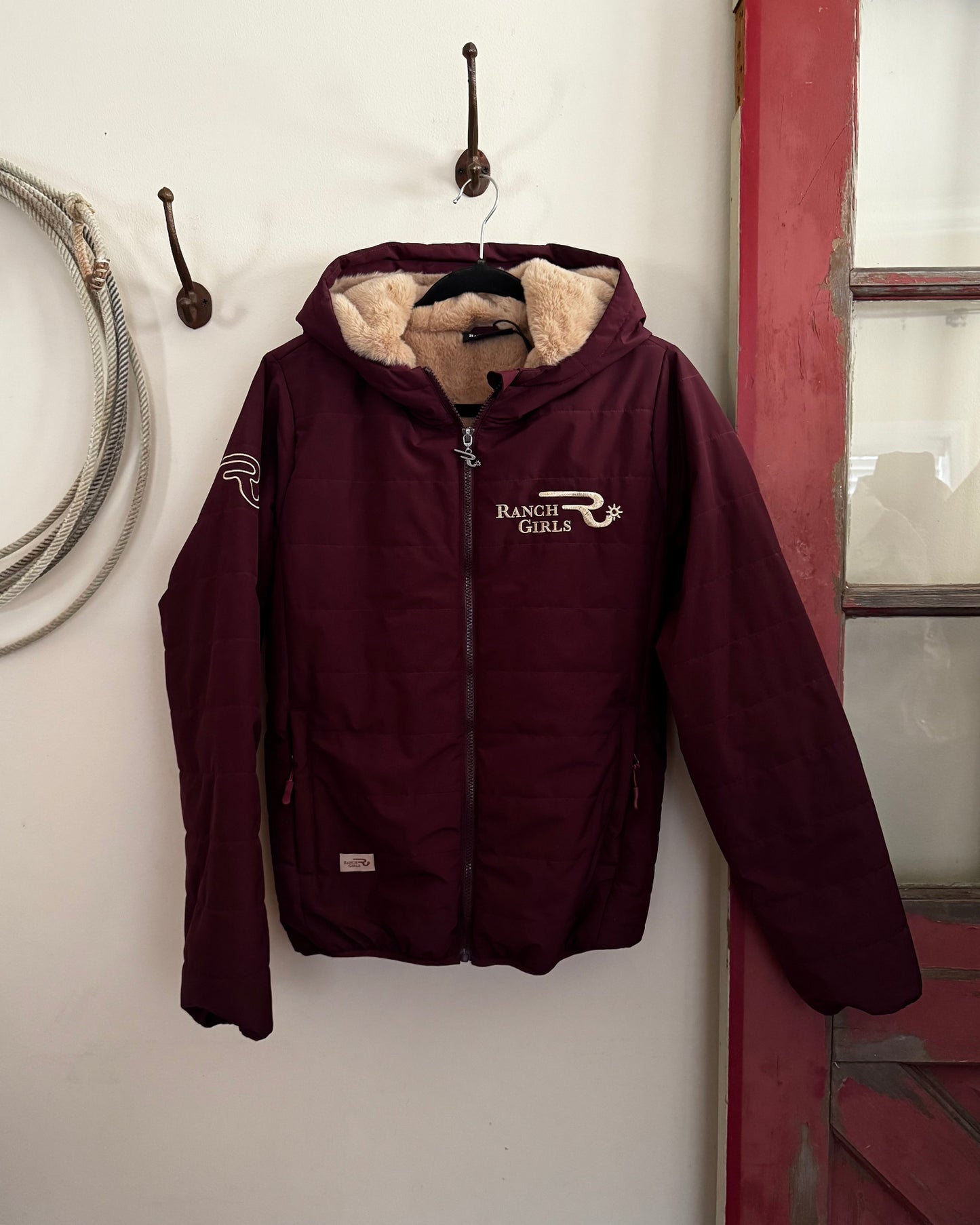 Maroon jacket with fur-lined hood hanging on a hook against a white wall.