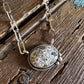 A silver-toned chain necklace with an oval pendant featuring a textured, crackled stone in cream, beige, and gray tones, displayed on a rustic wooden surface with a white faux-fur accent.