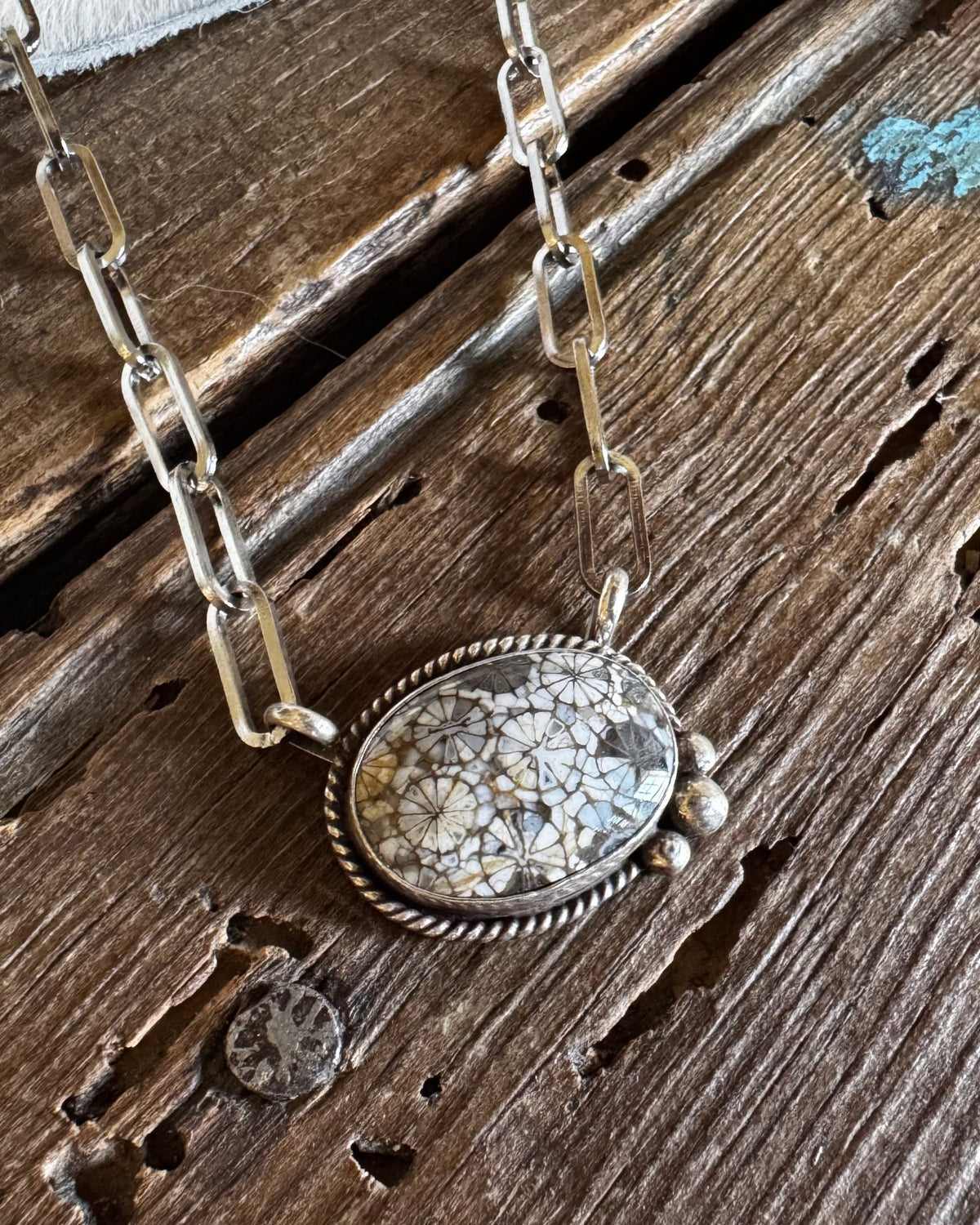 A silver-toned chain necklace with an oval pendant featuring a textured, crackled stone in cream, beige, and gray tones, displayed on a rustic wooden surface with a white faux-fur accent.
