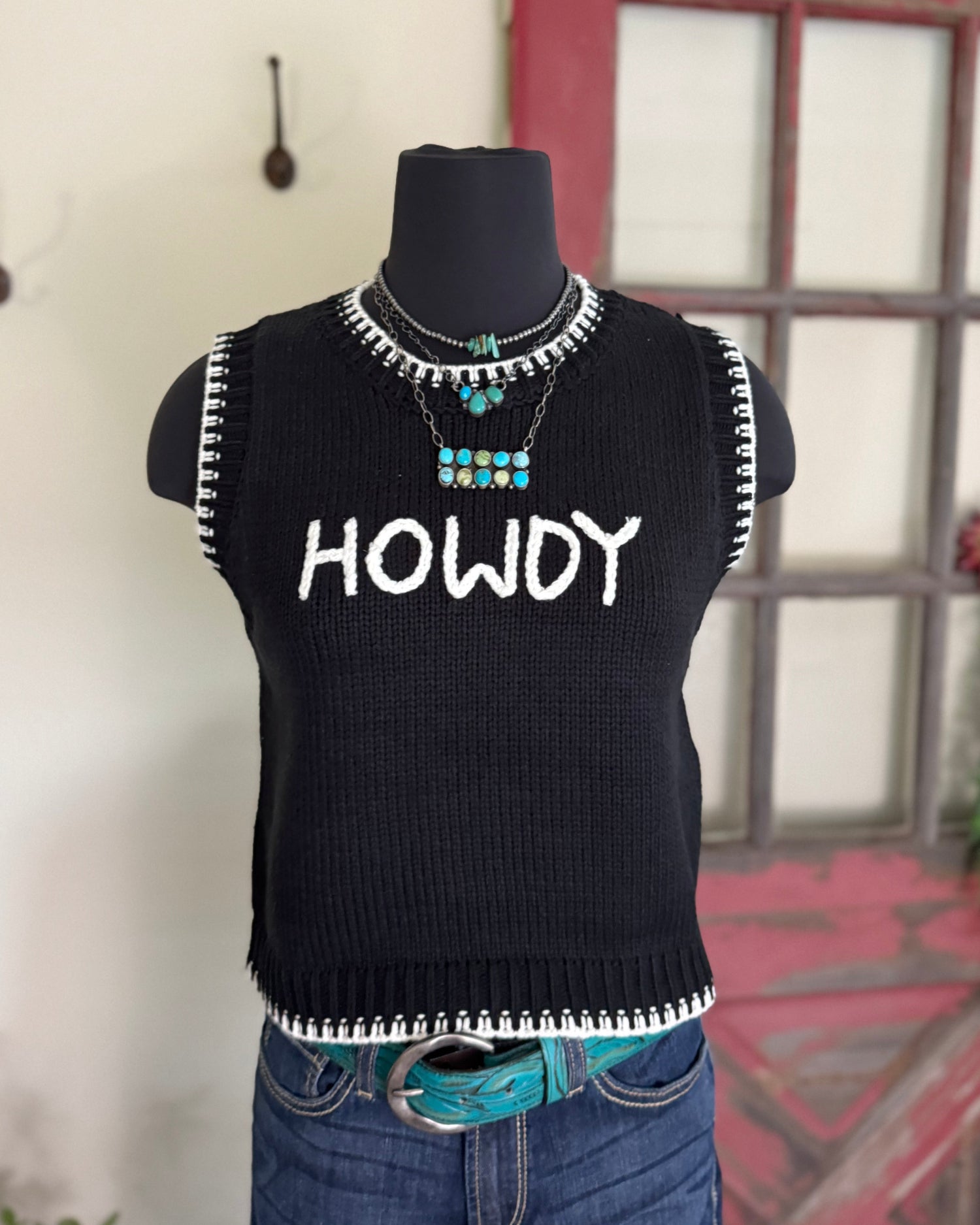 Black sleeveless sweater with 'HOWDY' text on a mannequin against a rustic background.
