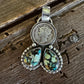 A silver pendant featuring a circular coin medallion and two teardrop-shaped turquoise stones, displayed on a rustic dark wood background.