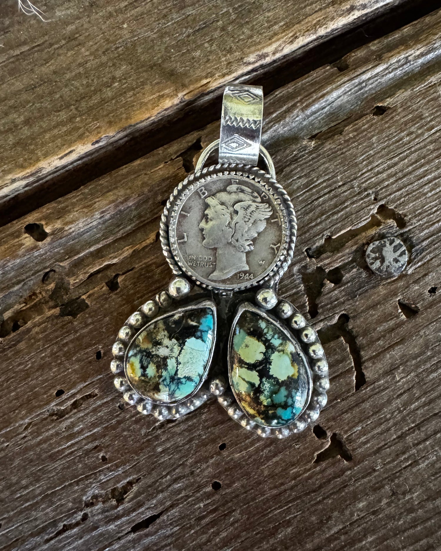 A silver pendant featuring a circular coin medallion and two teardrop-shaped turquoise stones, displayed on a rustic dark wood background.
