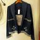 A dark navy blue knit cardigan with a shawl collar, featuring a textured geometric pattern on the chest and star motifs near the hem, hanging on a wooden hanger against a cream-colored wall.