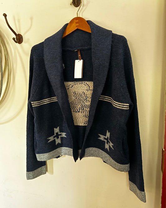 A dark navy blue knit cardigan with a shawl collar, featuring a textured geometric pattern on the chest and star motifs near the hem, hanging on a wooden hanger against a cream-colored wall.