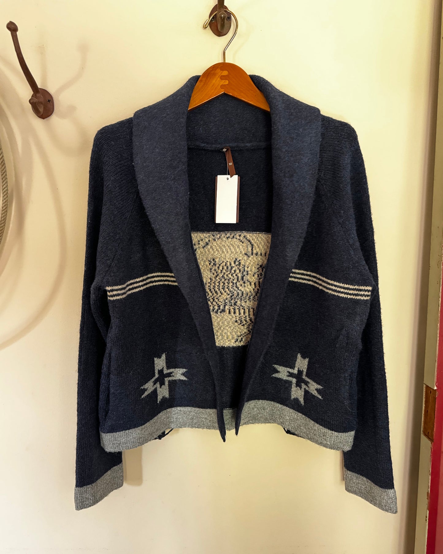 A dark navy blue knit cardigan with a shawl collar, featuring geometric star patterns near the hem and striped detailing on the chest, hanging on a wooden hanger against a cream-colored wall.