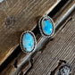 A pair of silver-toned oval stud earrings with light blue turquoise stones, displayed on a rustic dark wood surface.