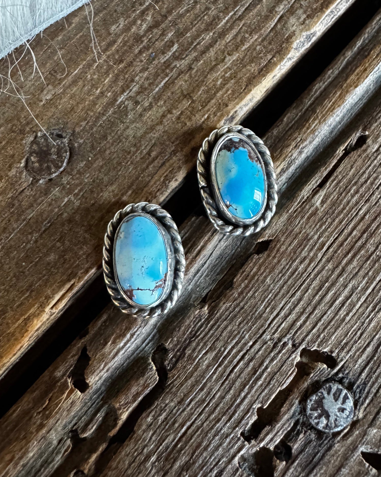 A pair of silver-toned oval stud earrings with light blue turquoise stones, displayed on a rustic dark wood surface.