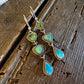 A pair of dangling earrings with triangular green and turquoise-blue stones set in silver-toned metal, displayed on a rustic dark wood surface.