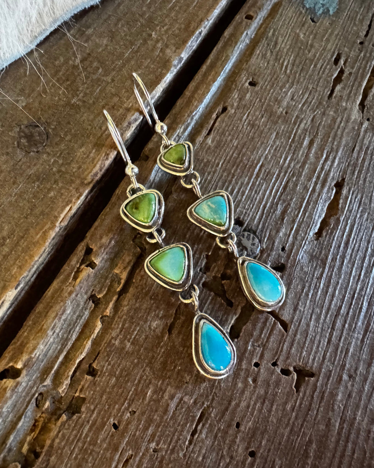 A pair of dangling earrings with triangular green and turquoise-blue stones set in silver-toned metal, displayed on a rustic dark wood surface.