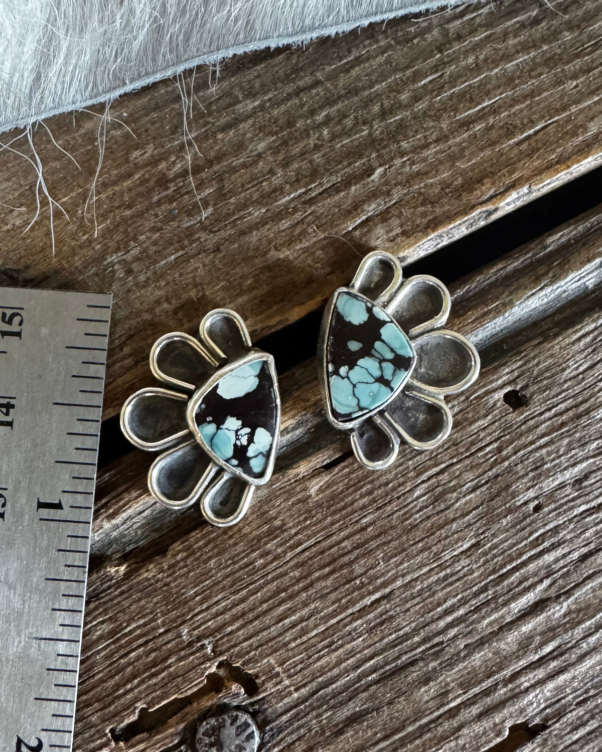 A pair of silver-toned stud earrings with triangular turquoise-blue and black marbled stone centers, displayed on a rustic wooden surface next to a metal ruler for scale.