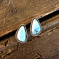 A pair of teardrop-shaped turquoise stud earrings with silver bezel settings, resting on a rustic dark wood surface.