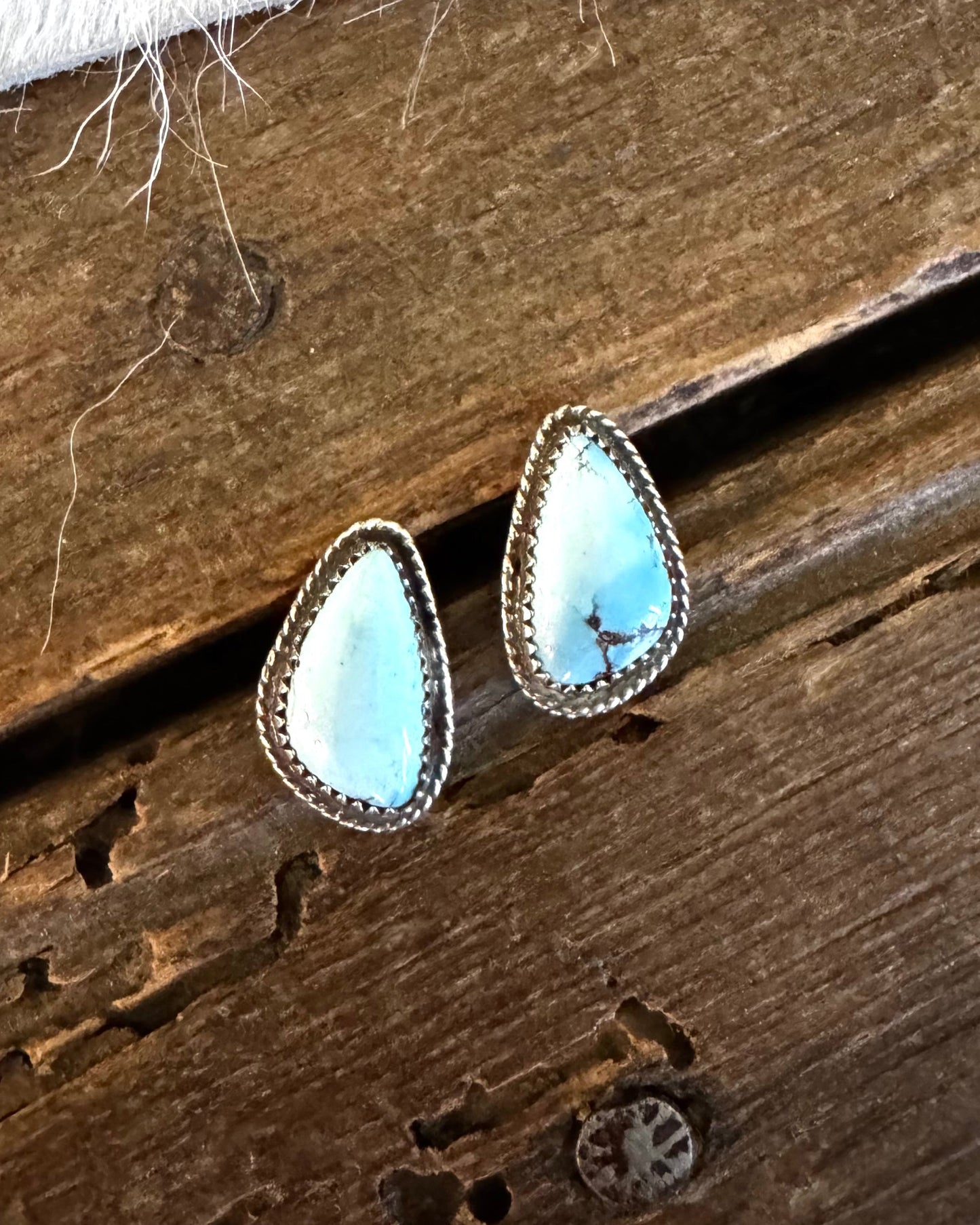 A pair of teardrop-shaped turquoise stud earrings with silver bezel settings, resting on a rustic dark wood surface.