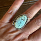 A close-up of a hand wearing a silver ring with an oval turquoise stone, resting on a dark wooden surface.