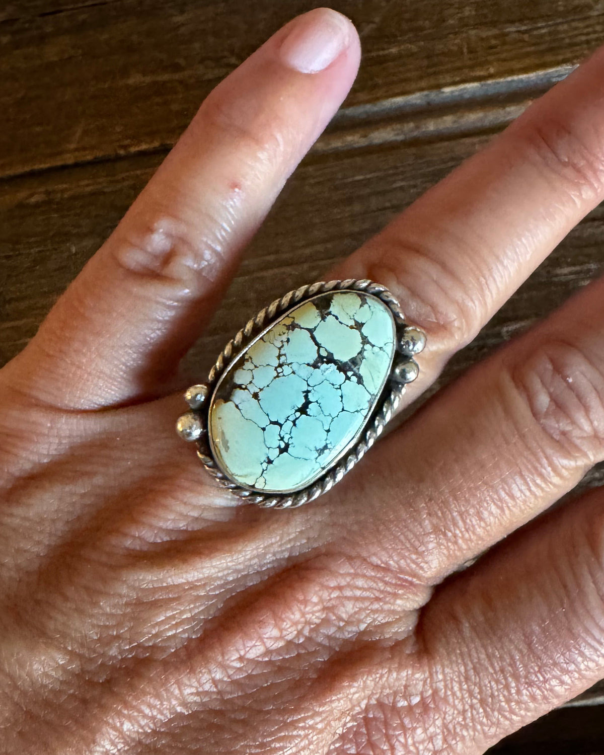 A close-up of a hand wearing a silver ring with an oval turquoise stone, resting on a dark wooden surface.