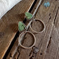 A pair of bohemian-style drop earrings with green stone triangles and twisted silver rings, displayed on a rustic wooden surface next to a white faux fur trim.