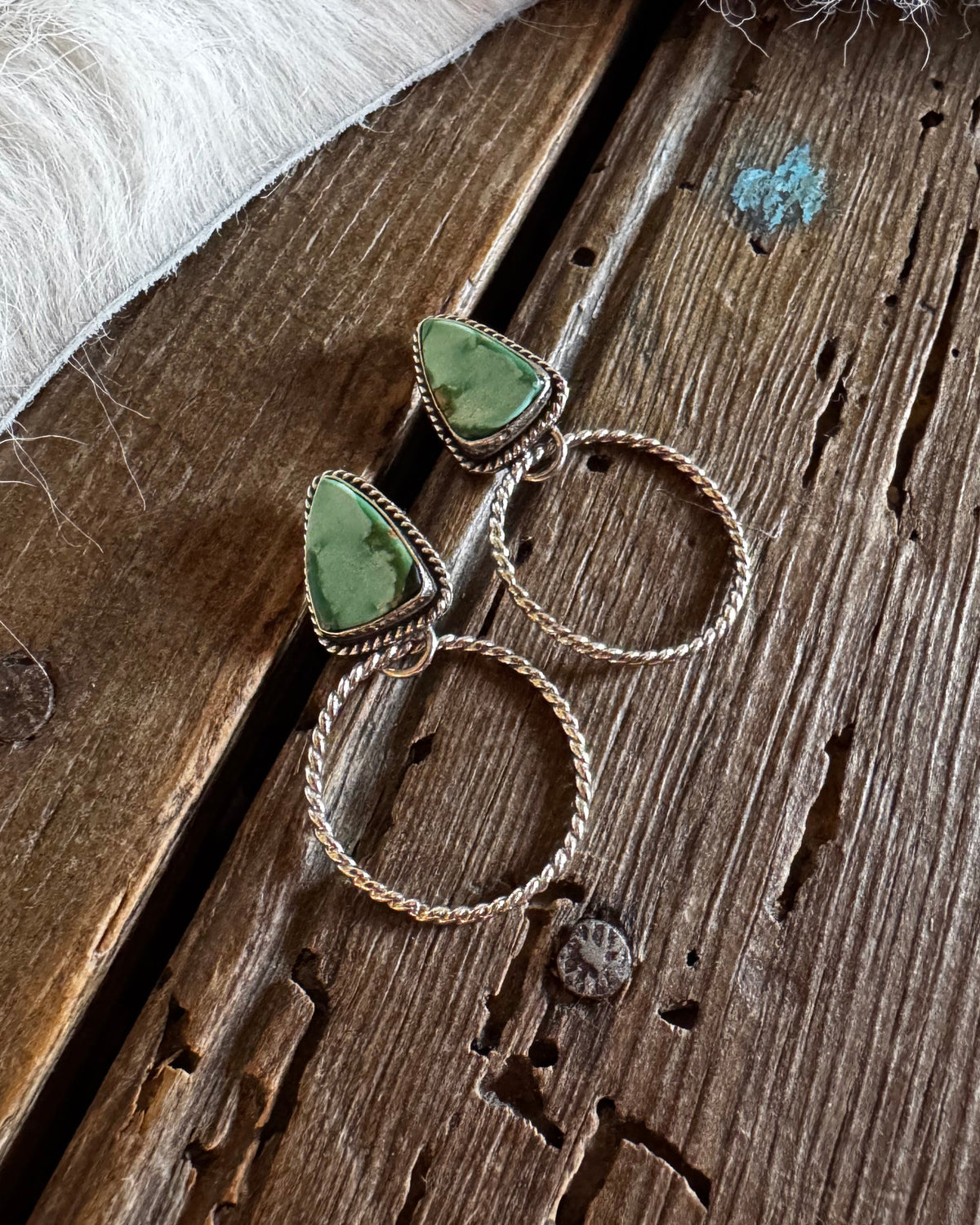 A pair of bohemian-style drop earrings with green stone triangles and twisted silver rings, displayed on a rustic wooden surface next to a white faux fur trim.