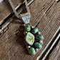 A silver-toned paw-shaped pendant necklace with green and white marbled turquoise-like stones, displayed on a rustic wooden surface with a white faux-fur prop.